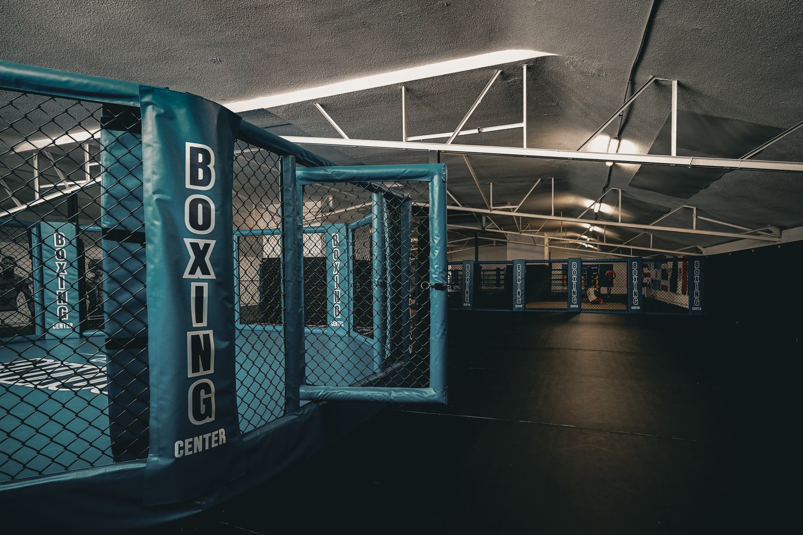 Un poteau de la cage MMA marqué Boxing Center et, derrière, toute la longueur de la grande salle du Boxing Center Toulouse États-Unis, vide