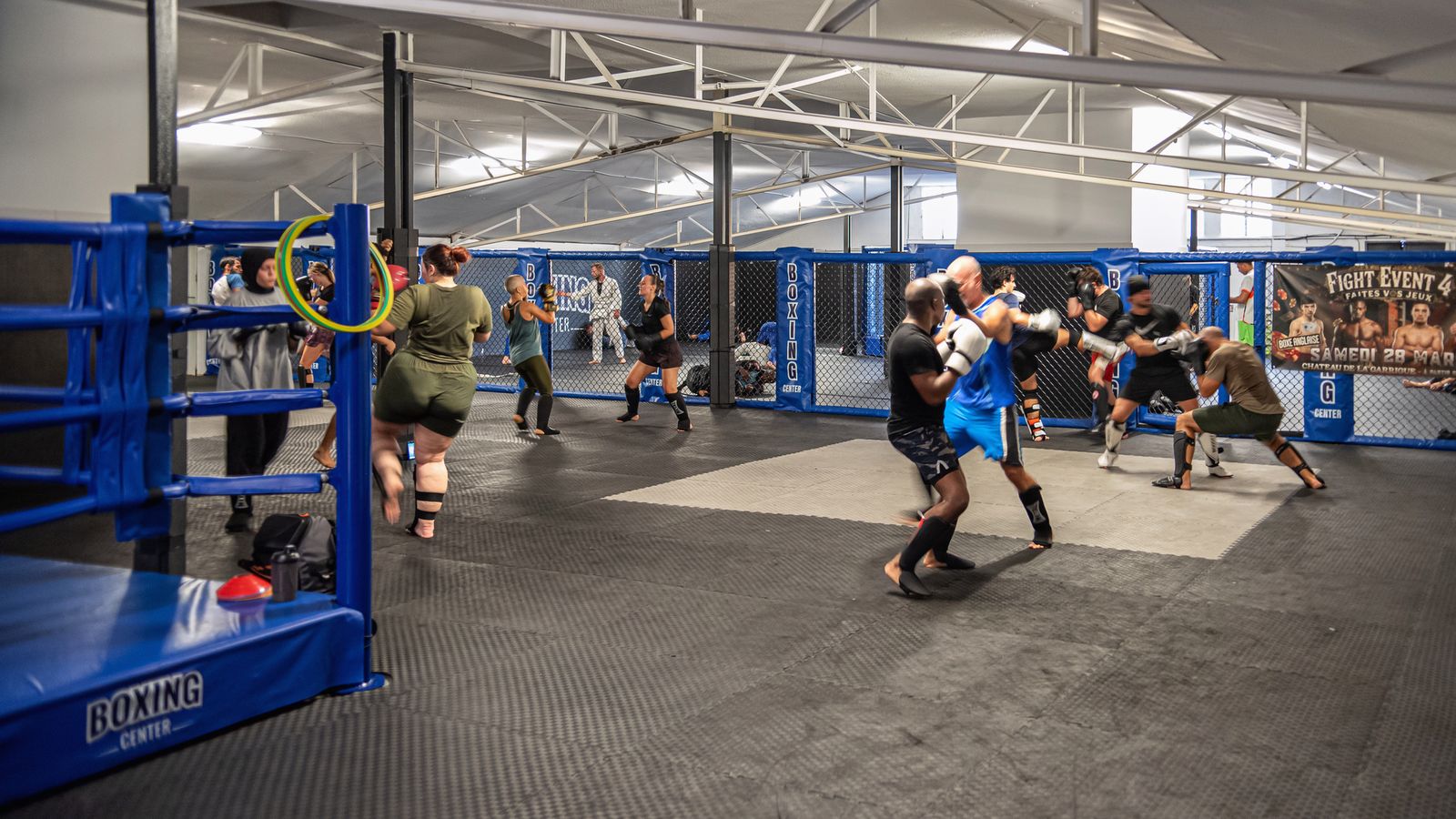 La grande salle : le ring au premier plan, la cage MMA au fond, un cours en pleine séance